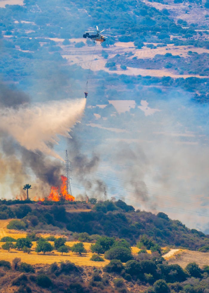 Aerial view of helicopter dousing forest fire with water bucket, showcasing fire control efforts.
